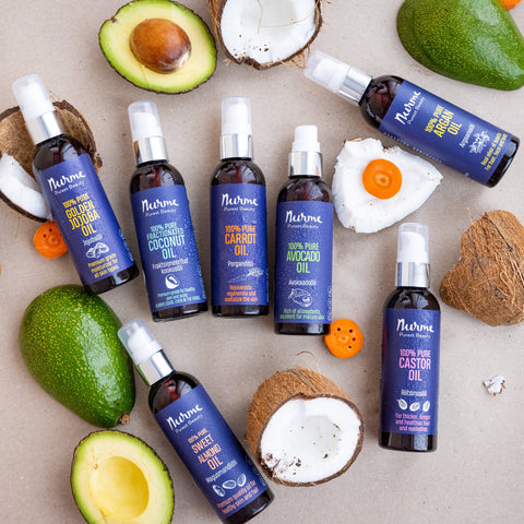 A selection of Nurme natural oils in dark bottles, including Golden Jojoba Oil, Fractionated Coconut Oil, Carrot Oil, Avocado Oil, Sweet Almond Oil, Argan Oil, and Castor Oil, surrounded by fresh avocado, coconut, and carrot pieces.