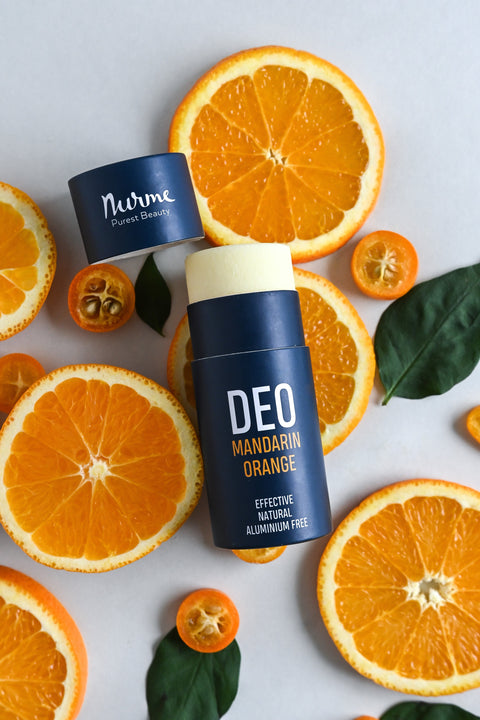 Nurme deodorants in blue packaging, featuring scents like Lemon Eucalyptus, Ylang-Ylang Bergamot, Mandarin Orange, Spearmint Rosemary, Lime Patchouli, and Lavender Sage, arranged on stones with a branch in the background.