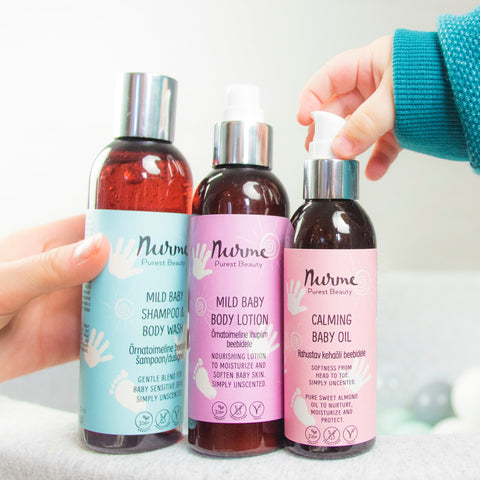 Nurme organic baby care products, including mild baby shampoo & body wash, mild baby body lotion, and calming baby oil, held by adult and baby hands on a soft fabric background.