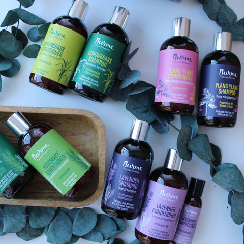 Hair care collection featuring lemongrass, rosemary, lavender, and ylang-ylang shampoos and conditioners, displayed with eucalyptus leaves and a wooden tray on a white background.