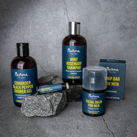 Men's grooming collection with shampoo, shower gel, face balm, lip balm, and soap bar in blue and yellow packaging, displayed on a concrete background with natural stones.